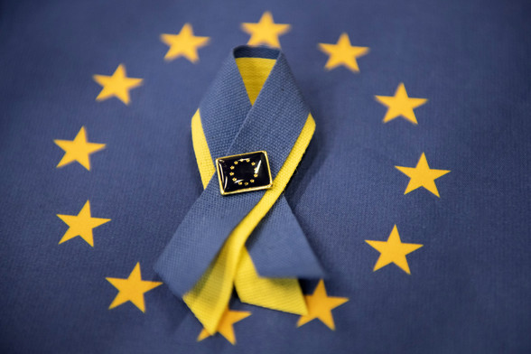 A photo of a ribbon with Ukrainian colours. The ribbon lies in the middle of the stars of an EU flag and has an EU pin on it.  